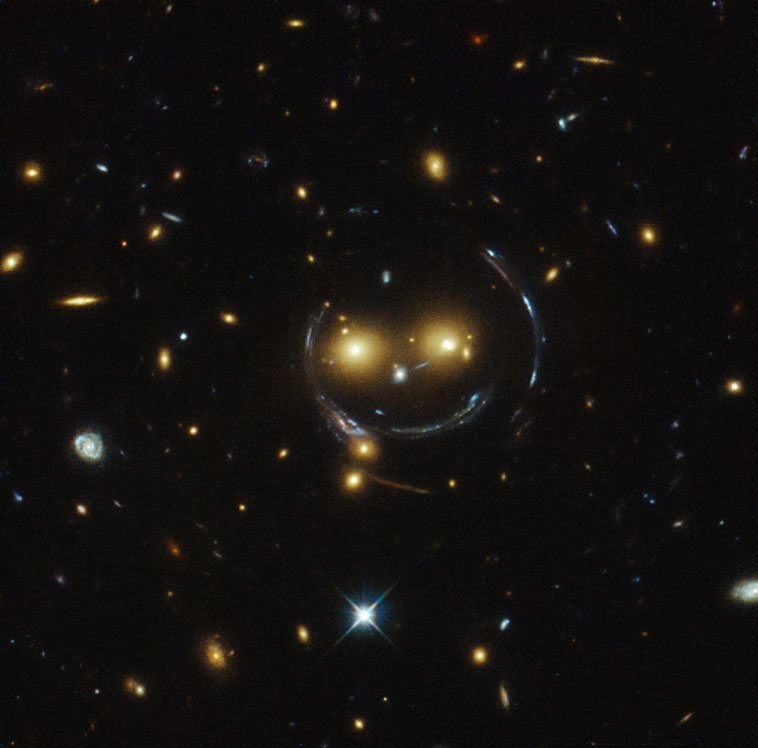 ハッブル宇宙望遠鏡が捉えた宇宙の「笑顔」。2つの銀河が「目」となり、強い重力レンズ効果で生じた円弧が「輪郭」と「口」を形作っている。