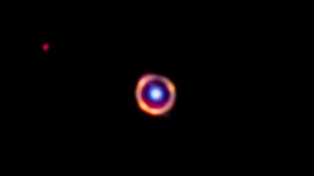 銀河系から120億光年以上離れた場所にある銀河SPT0418-47。はるか前方にある別の銀河の重力レンズ効果でリング状に見えている。青色が前方銀河、赤色が後方銀河、オレンジ色が今回検出された多環芳香族炭化水素（PAH）を示す。